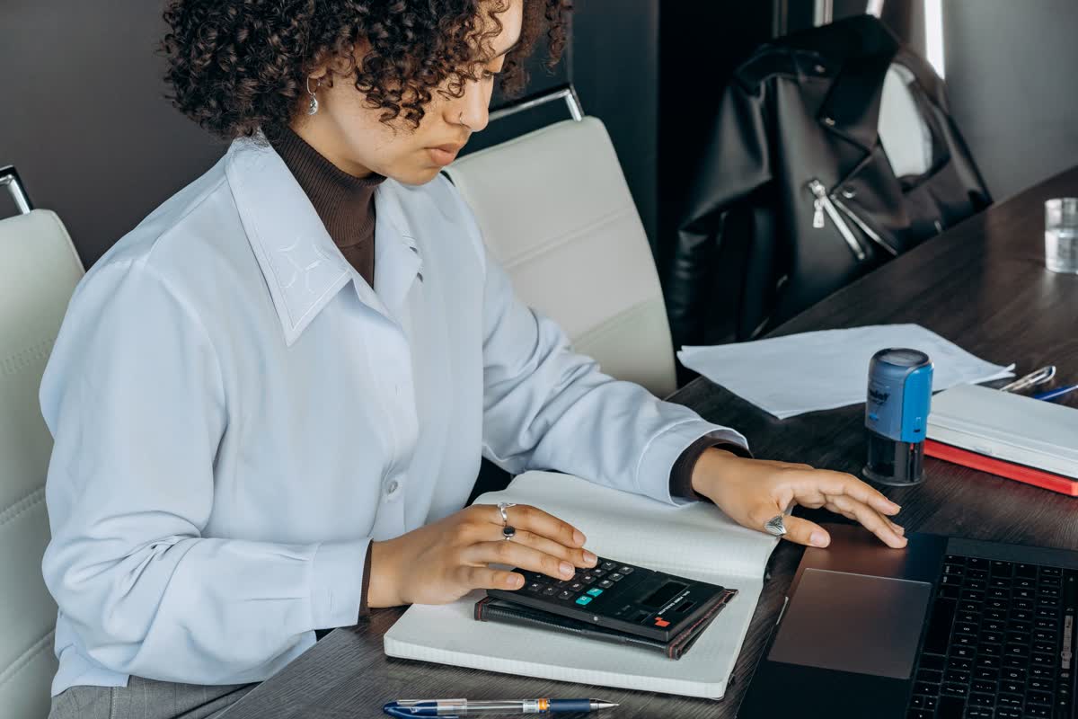 A person at a desk using a calculator and an open notebook beside a laptop and papers: an illustrative image for finance close work.