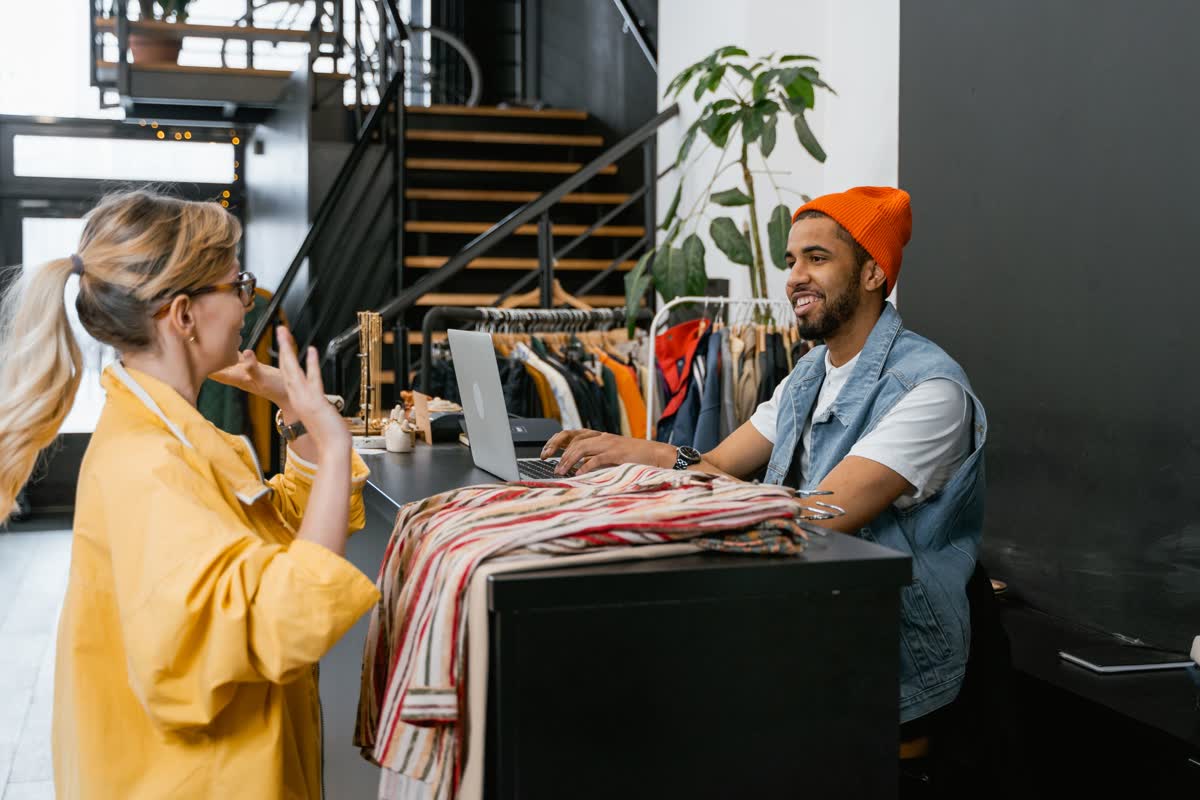 Two people talking across a counter in a clothing boutique, one at a laptop: an illustrative image for a sales-floor exchange.