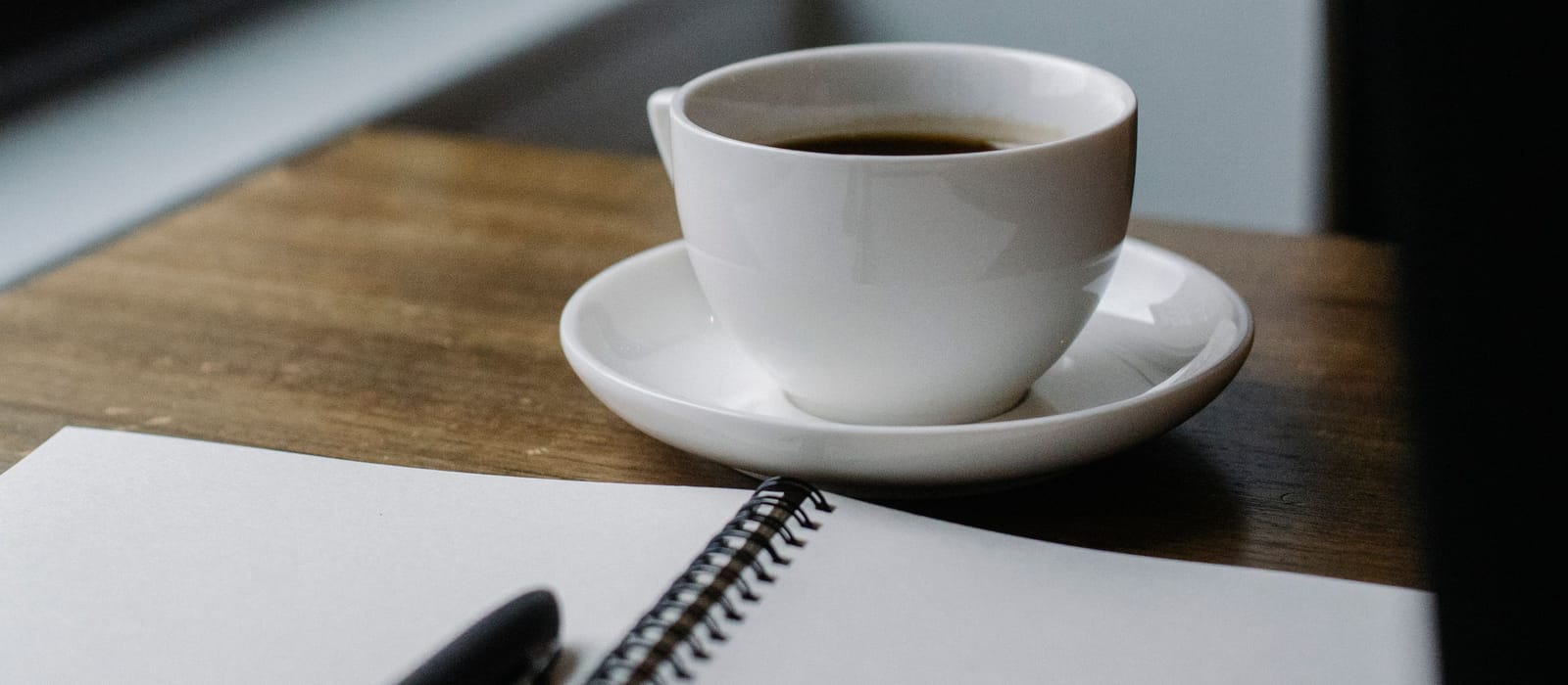 A coffee cup, a blank spiral notebook and a pen on a table by a window: an illustrative image for working a number out yourself.