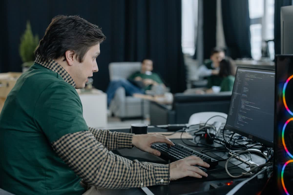 A person typing at a multi-monitor desk showing code, with colleagues seated in the background: an illustrative image for technology and IT work.